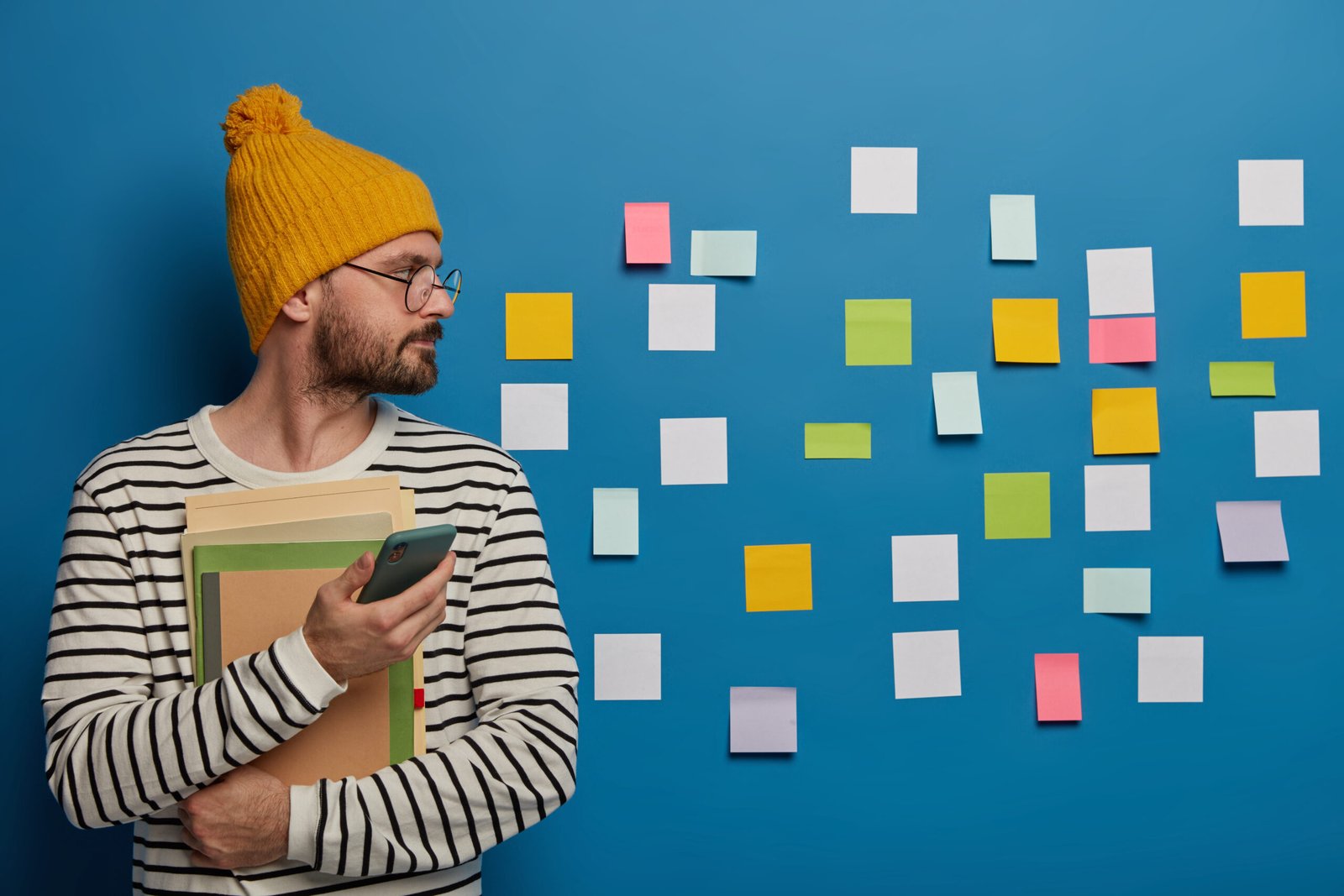 Indoor view of serious bearded man wears yellow hat and striped jumper, focused aside on wall with sticky notes, uses mobile phone, checks newsfeed, learns new information, holds notepad, books